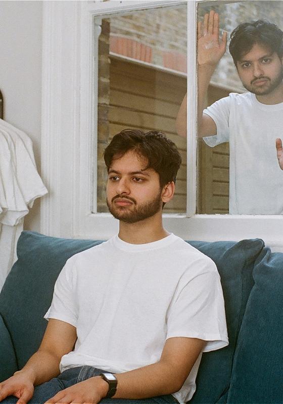 Rohan Sharma in a white t-shirt, sits on a couch. Several other white t-shirts hand on a rack next to him. in the window behind him, Rohan Sharma in a white tshirt is trying to break in