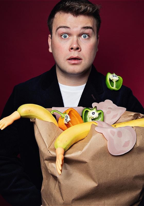 Max holds an overflowing bag of groceries, but the bananas have arms for some reason