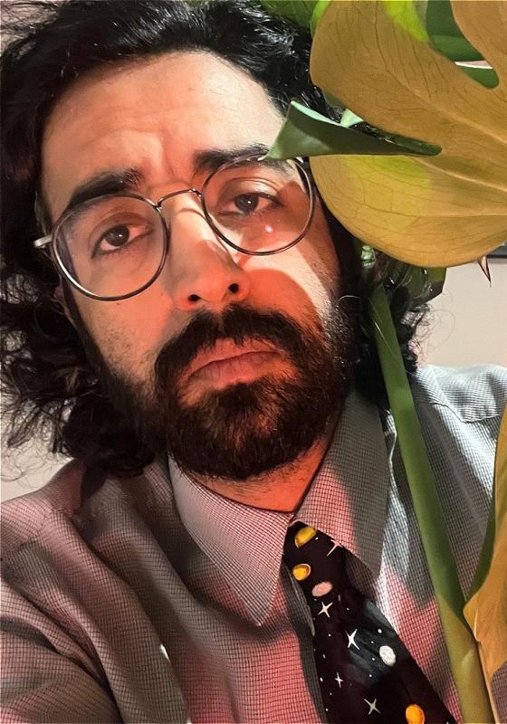 Rajiv is sad, in a tie, near some leafy plants. 