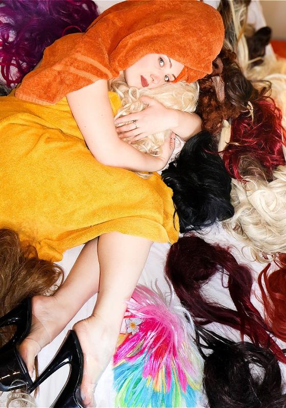 Alice lies on the floor in a yellow dress, surrounded by wigs.