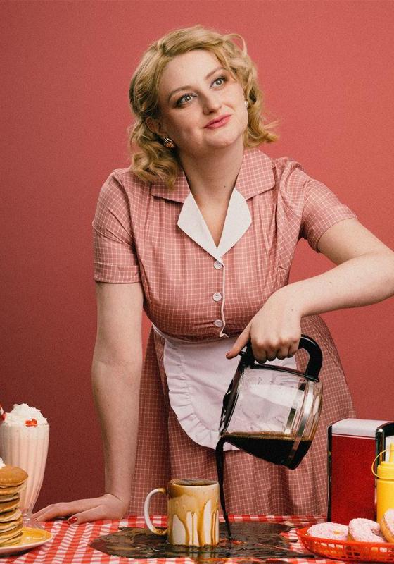 Lorna, wearing a pink diner dress, pours coffee into a mug, but misses. 