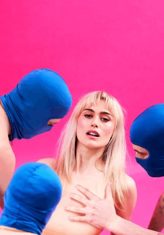 A blonde woman looks at the camera as three men in blue masks look at her