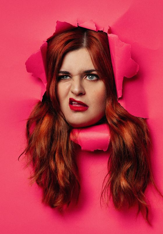 Alex's disgruntled face, framed by auburn hair, bursts through a hot pink paper.