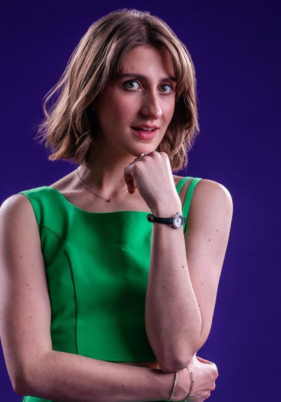 Anna Hale in a green dress on a purple background, resting her head on her hand. 