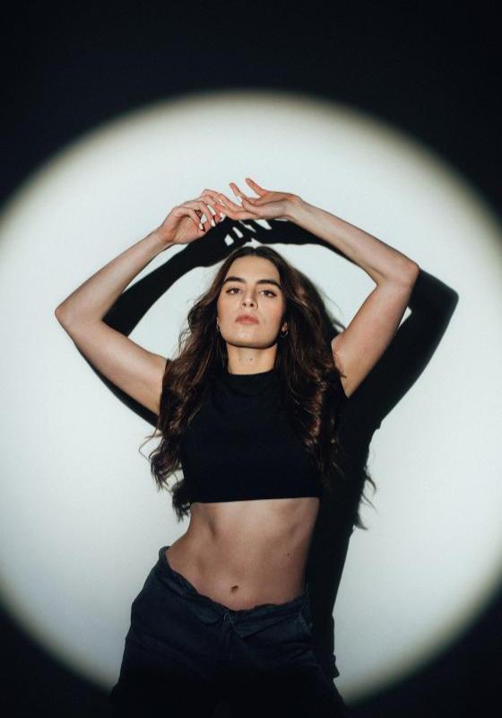 Amelia poses with her arms above her head in a black crop top - there is a front spotlight on her