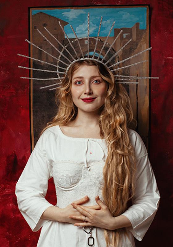 Bebe Cave wearing a long white dress and a headpiece gazes submissively into the camera.