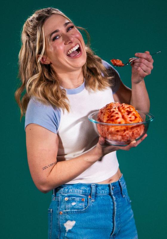 A woman in a t-shirt and jeans eating from a bowl containing a brain covered in sauce.