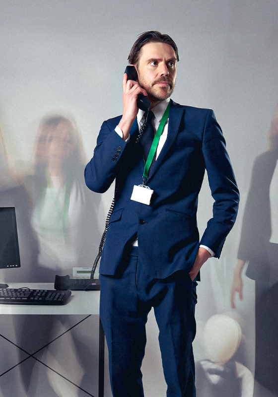 Ben in a blue suit, near an office desk, on the phone, while blurry office people move behind him