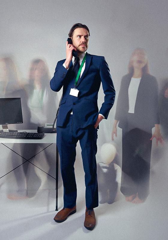 An actor wearing a suit and lanyard holding a phone with his hands in his pockets. Other characters in the background are blurred out.