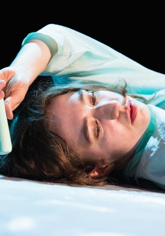 The image is a shot of the performer’s face against a dark background. The performer is white afab and has their eyes downcast. The performer is lying on the floor with their left arm reaching over the top of their head and their left hand holding a piece of light blue chalk. This image symbolises a moment in the show when the performer is drawing a chalk circle around themselves creating a physical outline of the body connected to how trauma is held in the body.