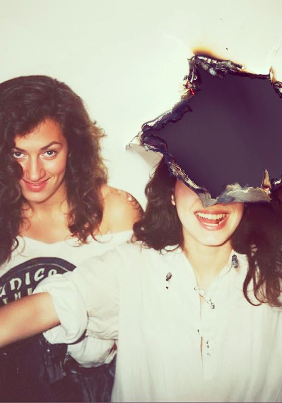 Two people standing together in white tops, smiling, one with their face covered. 