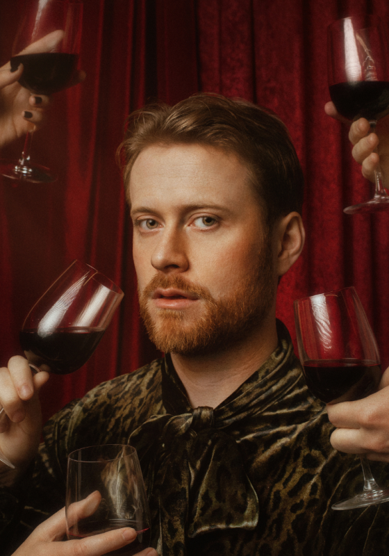 John's head, framed by five hands holding glasses of red wine, is seen against a red drape.  