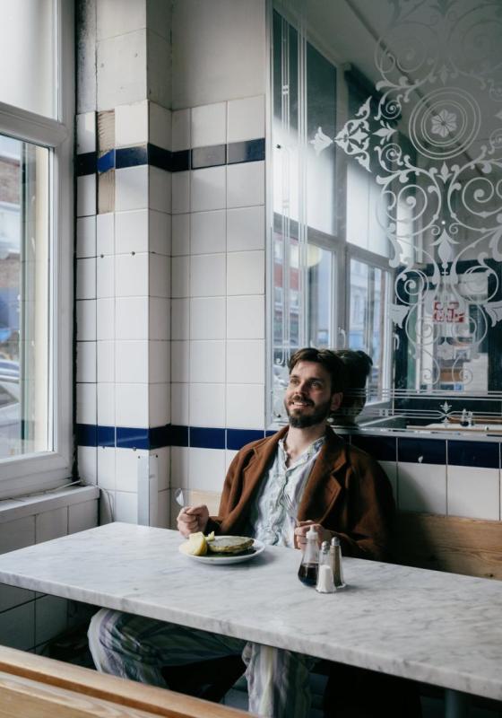 Joz sits in a no frills cafe wearing pyjamas and a loose brown overcoat.
