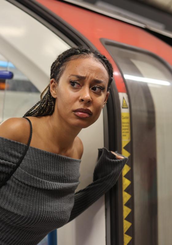 Performer wearing a jumper leans outside of an open london underground carriage looking wary.
