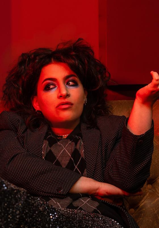 Leila sits on a worn leather sofa with arms crossed holding up a bottle of pills.