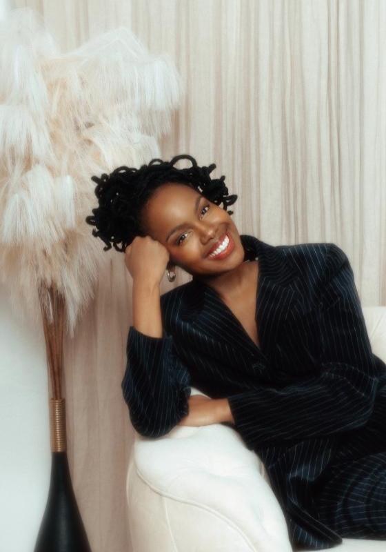 Sharon in a fantastic black suit, sits on a white couch with white drapes behind her, leaning her head on her hand. 