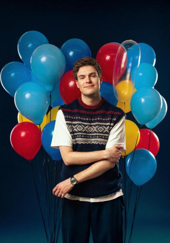Thor stands in front of a big bunch of balloons wearing a knitted vest over a white t-shirt.