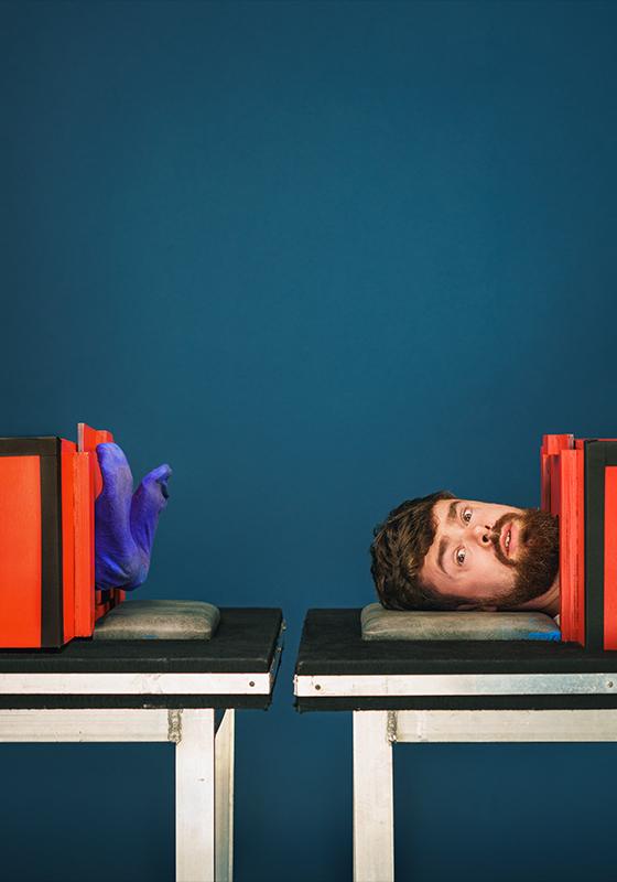 On the left a pair of blue socked feet poke out of a red sawing in half illusion. On the right Tom Brace's head pokes out of a red sawing in half illusion. Both are against a blue background.