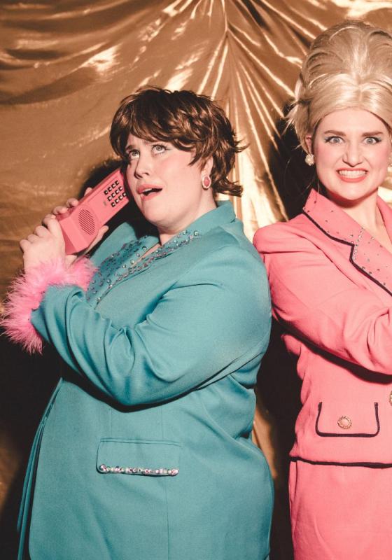 Sally and Susan, the shows protagonists, are smiling widely at the Camera. Susan has a blond high beehive, and is wearing a pink skirt suit. Sally has choppy brown hair and is wearing a teal skirt suit. They are both holding pink plastic phone