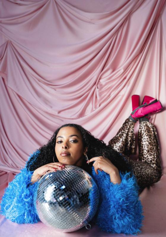 Performer lounges on top of a disco ball in a blue feather cardigan, leopard print leggings and heels.