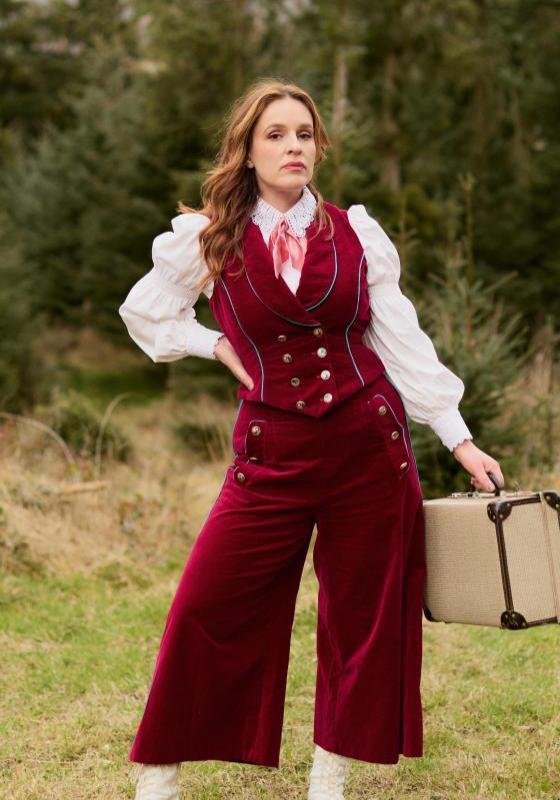 Katie stands in a field with a forest behind her, in a red velvet western inspired outfit, holding a vintage suitcase