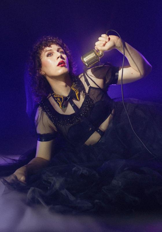 Sam lounging elegantly on the floor with a microphone wearing a black mesh ballgown with a smokey purple haze