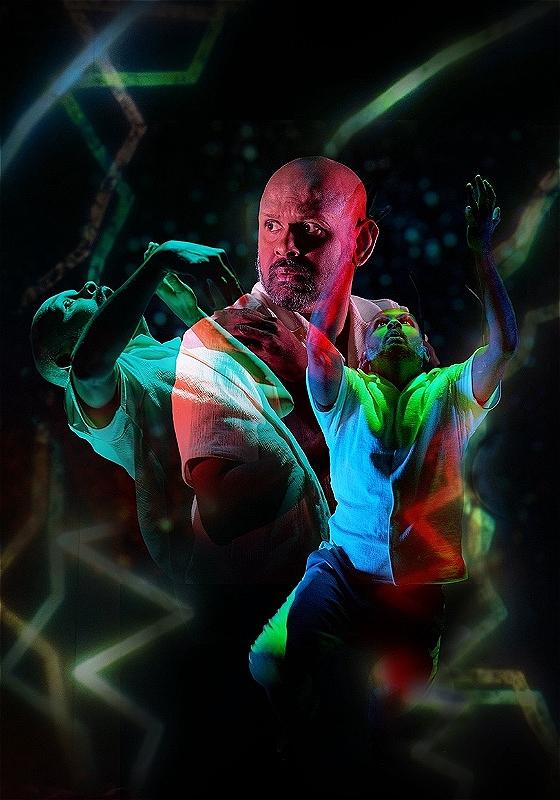 Ethereal image of a stocky bald man looking pensively over his shoulder whilst two figures dance expressively. The three men all blend together with green and red lights.