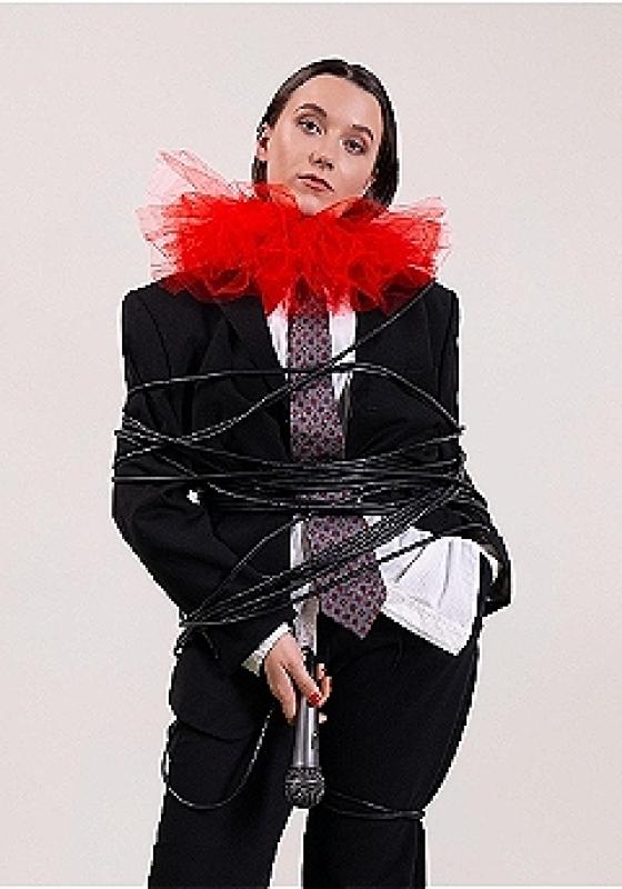 Claire Noy stands in a suit, tie and large red ruff looking particularly apathetic about the fact somebody has wrapped their microphone cable around them twenty times. They hold the microphone laxly in their right hand.