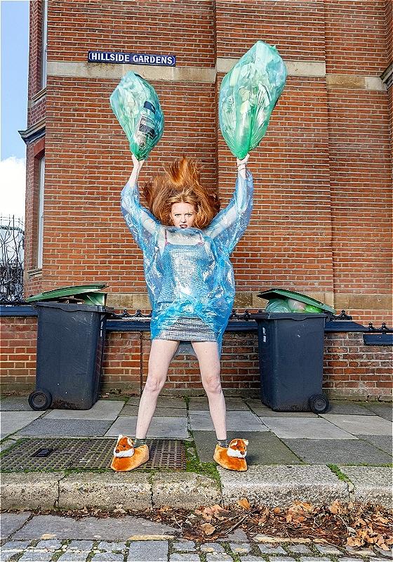 A red haired lady looks confused at the camera with a big blue recycling bag draped across her body. In her hands she hoists two full green recycling bags above her head in a menacing position.