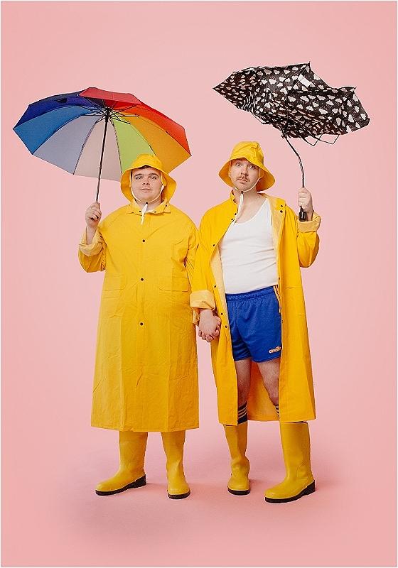 Two performers with hands joined are holding up umbrellas wearing yellow coats and matching fisherman hats.
