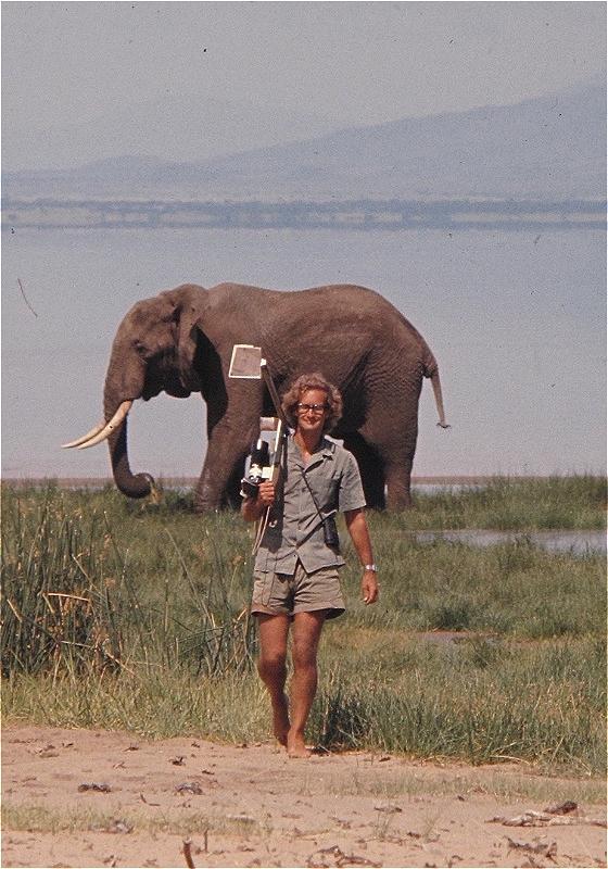 Iain Douglas-Hamilton carries a camera and tripod away from a lake where an elephant is stood.