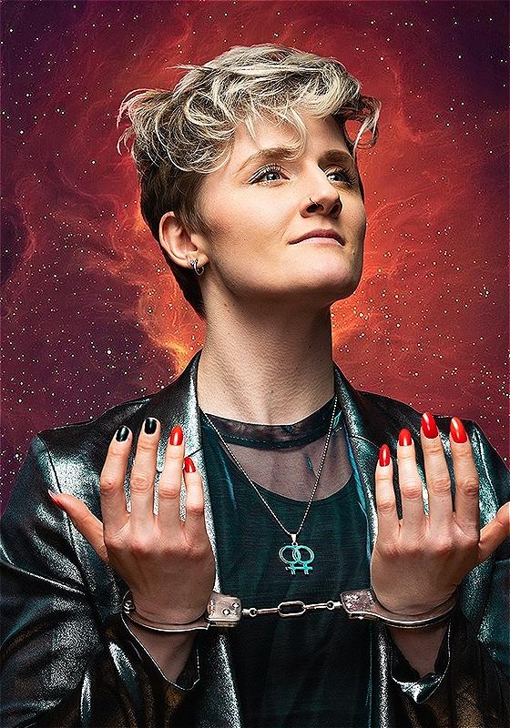 Performer stands against an intergalactic starry background with their hands in handcuffs.