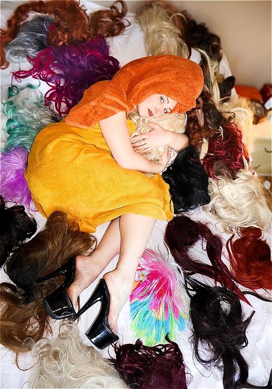 Alice lounges on a bed in the fetal position surrounded by a great many number of exuberant and vibrant wigs. She cradles one wig in her arms like a babe.