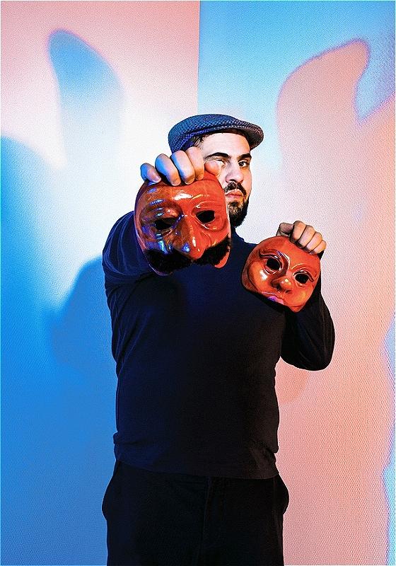 A performer wearing a cap holds up two theatre masks with one arm outstretched.