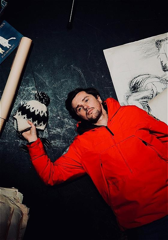 A man in a bright red jumper lays down next to line drawings, clutching a woolen trapper hat in his right hand