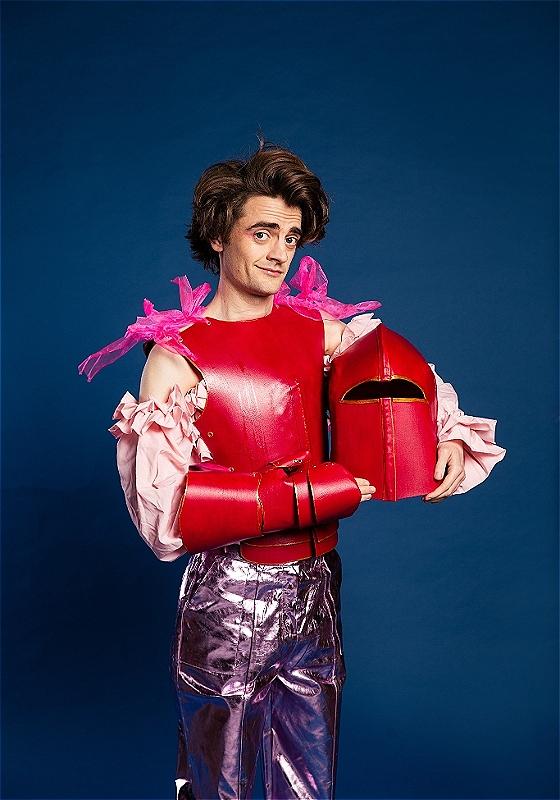 A man with very bouffant hair adorns a red DIY knights costume, holding the helmet and looking at the camera cheekily.