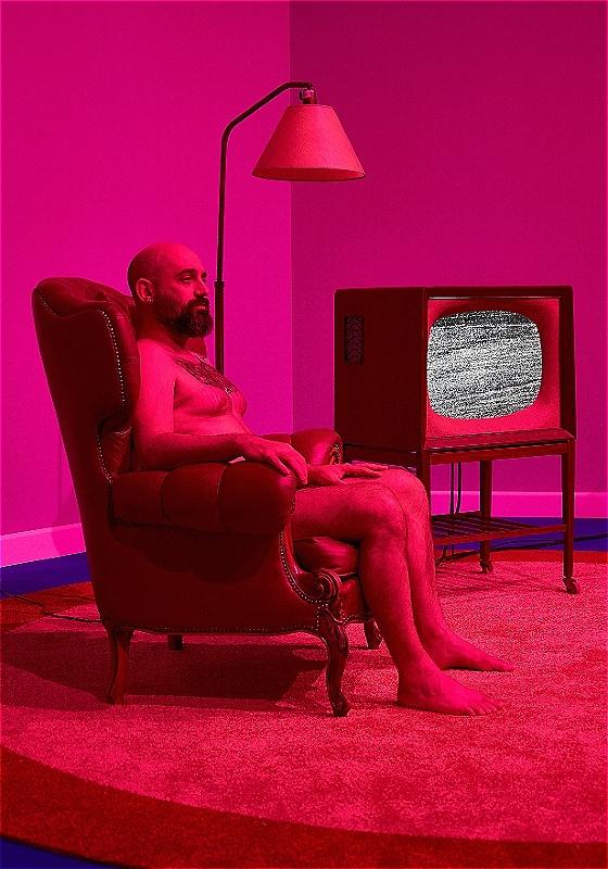 A man sits in a armchair next to a lamp and tv set with a heavy pink filter.