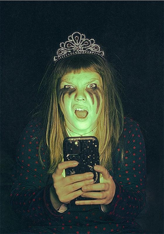 A ghastly ghoul lady stands with tiara on head looking perplexed by what she sees on her mobile phone which is planted in front of her face.