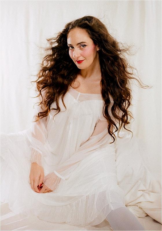 Cat Cohen sitting and smiling sweetly in an angelic white gown against a white fabric backdrop