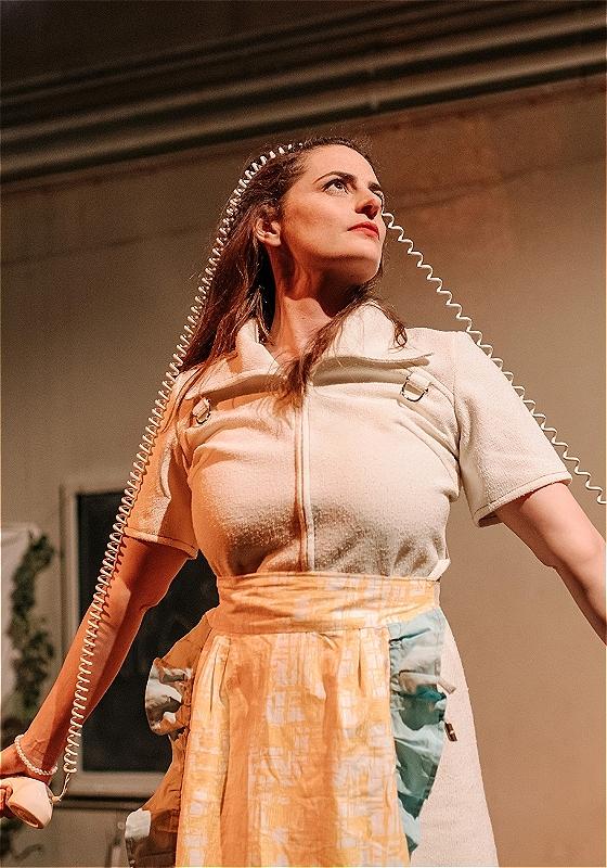 A performer with long brown hair stands up straight striking a power pose, with a corded telephone receiver in one hand and its main body in the other. The curly cord connecting them stretches over the performer's head.