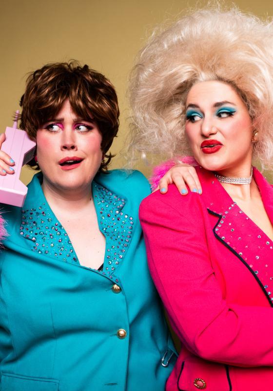 The performers in garish big blue and pink 80s style suits and wigs, one holding a vintage pink phone