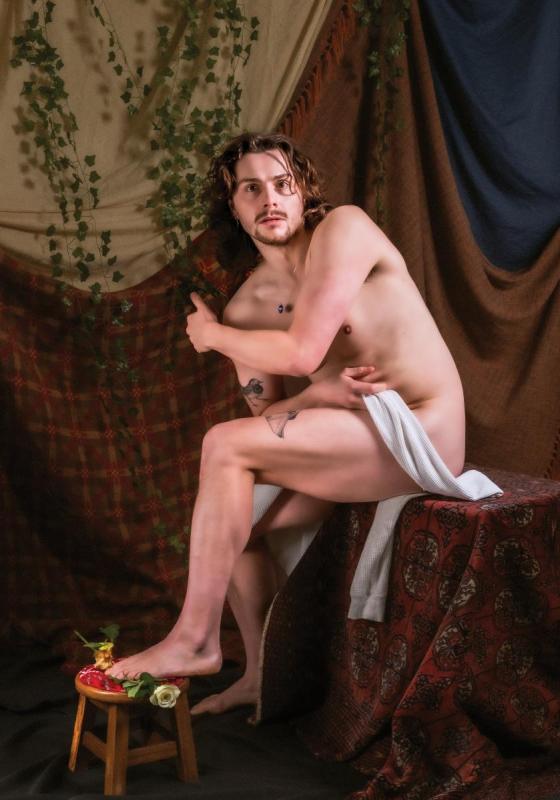 Sam, shirtless, stares into the distance behind the camera. There are vines and curtains draped in the background. 