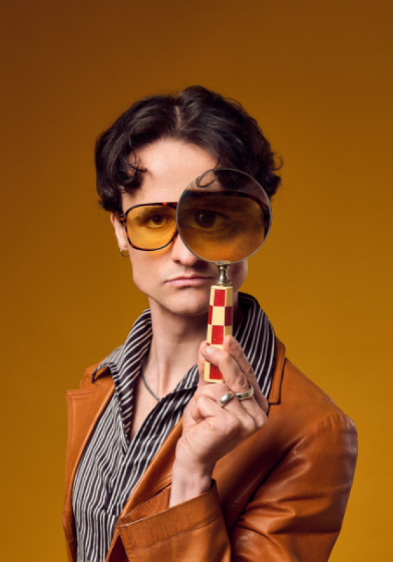 Jazz holding a magnifying glass up to his eyes wearing a 70s inspired outfit