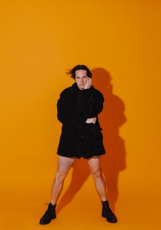 Luke Mcqueen wearing a long black raincoat and no trousers standing against a bright orange background