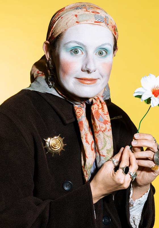 A person with white make-up wearing a headscarf, rings and holding a flower looks out at the viewer.