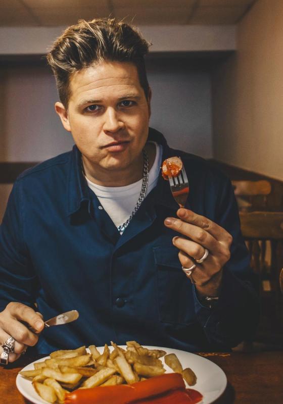 Luke Wright sits at a table of what looks like a greasy spoon type restaurant. They're holding up a fork that's got sausage and ketchup on it, and smiling at the camera with their mouth closed, mid-chew.