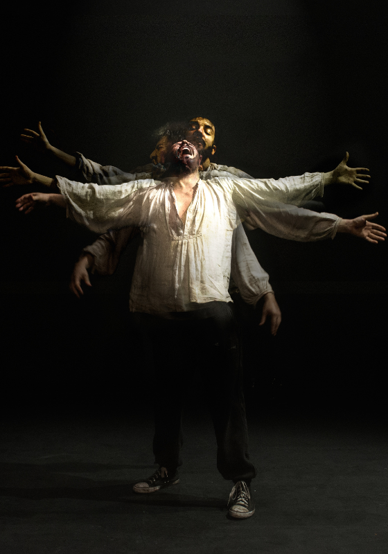 A performer in a loose white shirt stands with their arms outstretched, against a black background on an empty stage. They appear to be crying to the heavens. There are faint images of them in very slightly different poses superimposed on this main one. The effect means that it looks like they have six arms, and three heads, all doing something very slightly different.