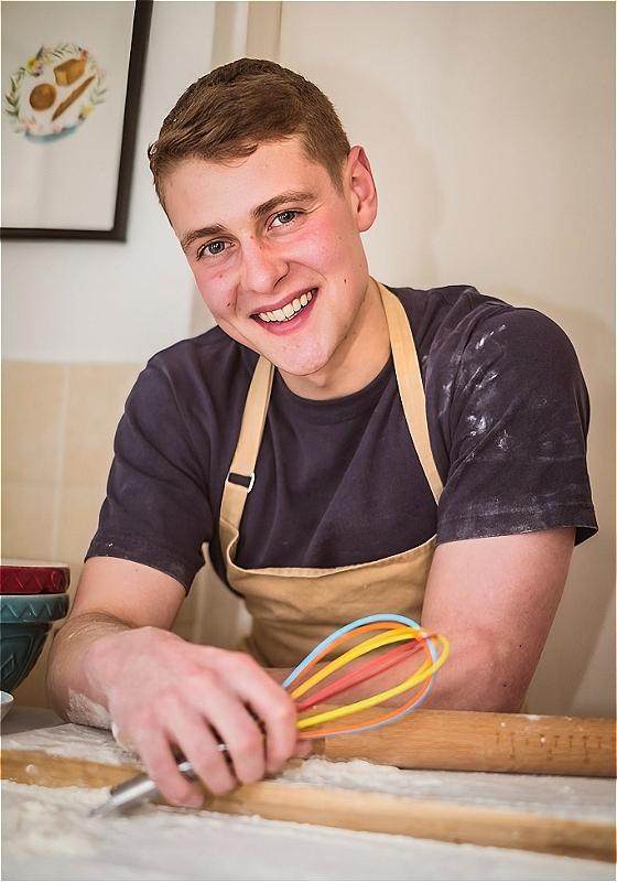 A smiling Peter covered in flour smiles at the camera holding a whisk.