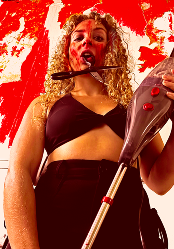 Performer with scissors hanging out of their mouth, holding a vacuum clear with blood over their face.