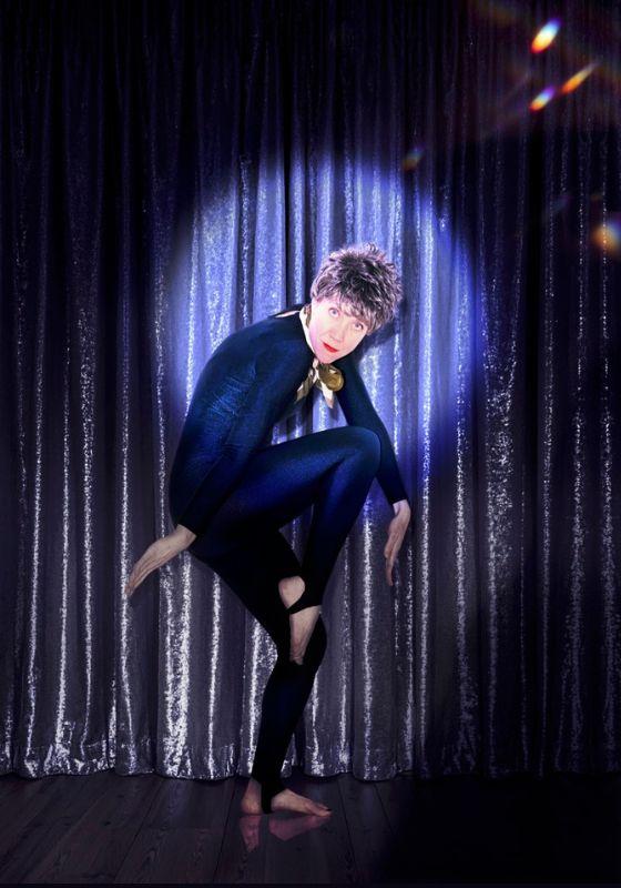Dusty, wearing a black leotard, dances freely against a dark blue curtain with a spotlight on their face